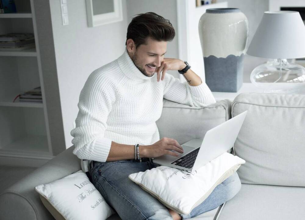 Junger Mann arbeitet am Laptop und sitzt auf einem gemütlichen Sofa im Apartment.
